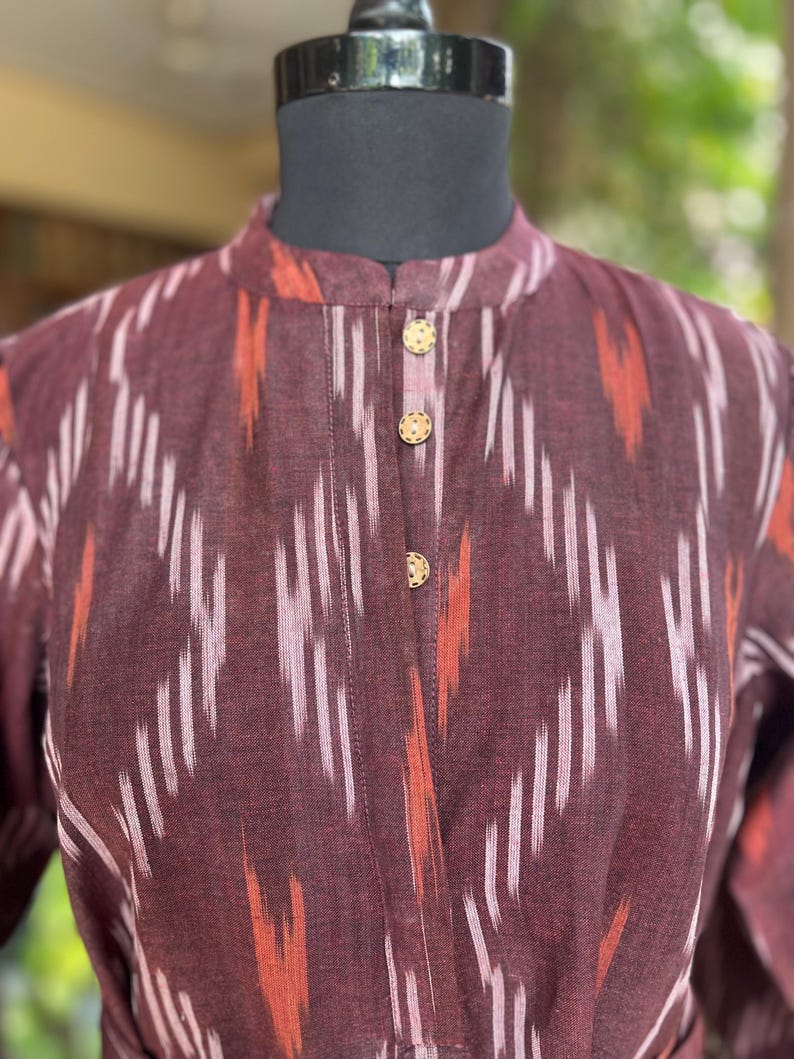 May include: A maroon dress with a mandarin collar and three gold buttons. The fabric features a white and orange ikat pattern. The dress is displayed on a black mannequin head.