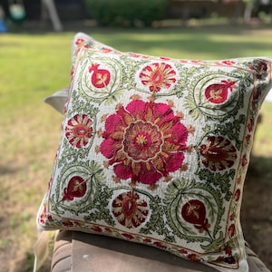 以下が含まれることがあります： クリーム色の背景に刺繍の花柄が施された正方形の装飾用クッション。デザインは、赤と金のトーンの大きな中央の花と、小さな赤い花と緑の葉で囲まれています。