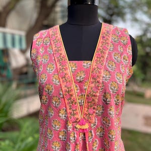 Puede incluir: Túnica rosa sin mangas con un estampado floral de flores amarillas y blancas. El escote está adornado con un borde floral y una borla amarilla. La túnica se exhibe en un maniquí negro.