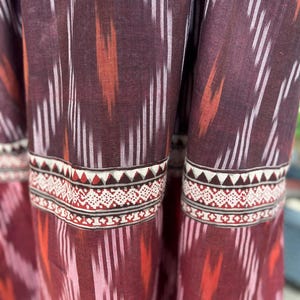 May include: Close-up of a fabric with a repeating pattern of vertical white and orange stripes against a deep burgundy background. The bottom edge features a decorative border with geometric shapes in red, white, and brown.