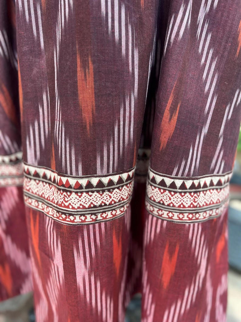 May include: Close-up of a fabric with a repeating pattern of vertical white and orange stripes against a deep burgundy background. The bottom edge features a decorative border with geometric shapes in red, white, and brown.