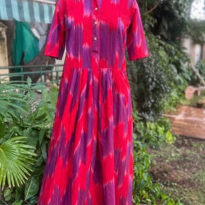 May include: A long, red and purple patterned dress with a collared neckline and short sleeves. The dress features a button-down front and a pleated skirt. The fabric has a vertical, ikat-style design. The dress is displayed on a mannequin.