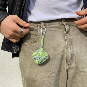 Könnte beinhalten: Kleine, quadratische Häkeltasche in Grün- und Blautönen, befestigt an einem silbernen Karabinerhaken. Der Haken ist am Bund einer khaki-farbenen Jeans befestigt. Eine schwarze Lederjacke und ein weißes T-Shirt sind sichtbar.