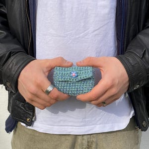 May include: A small, crocheted pouch in shades of blue and green, held in hands. The pouch has a flap closure with a pink and blue flower-shaped button. The item appears handmade and has a textured surface.