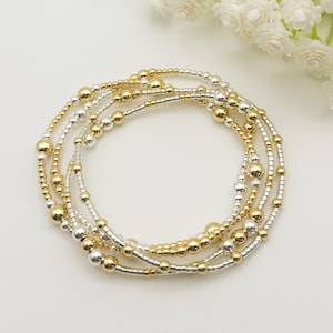 May include: A set of beaded bracelets in gold and silver tones. The bracelets are made with small and large beads, creating a layered look. The bracelets are displayed on a white surface with white flowers in the background.