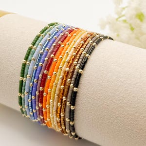 May include: A collection of colorful beaded bracelets displayed on a cream-colored surface. Each bracelet features small, vibrant beads in a different color, accented with gold-colored beads. The bracelets are arranged in a rainbow spectrum.