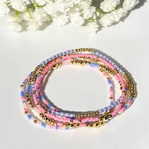 Puede incluir: Un conjunto de cinco pulseras de cuentas en rosa, azul y dorado. Las pulseras están apiladas una encima de la otra y están hechas de pequeñas cuentas redondas.