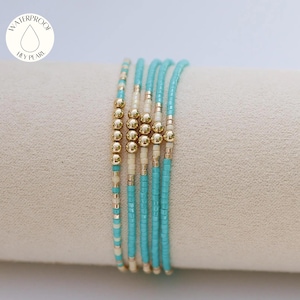 May include: A set of five beaded bracelets featuring turquoise and cream-colored seed beads accented with metallic gold-toned spherical beads. The bracelets are arranged in a row and include a circular logo with a water drop icon and the text WATERPROOF HEY PEARL.