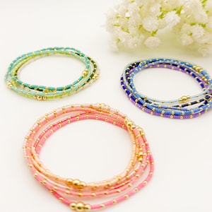 May include: Three beaded bracelets in shades of pink, blue, and green. The bracelets are made of small, colorful beads and gold-colored accents. The bracelets are arranged in a circular shape, with a sprig of white flowers in the background.