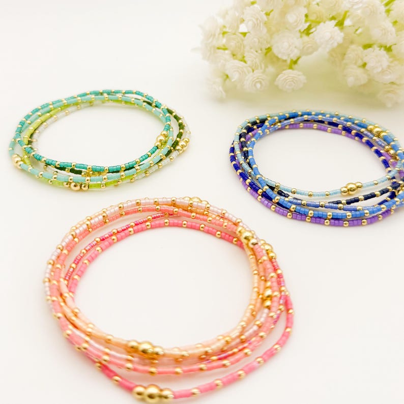 May include: Three beaded bracelets in shades of pink, blue, and green. The bracelets are made of small, colorful beads and gold-colored accents. The bracelets are arranged in a circular shape, with a sprig of white flowers in the background.