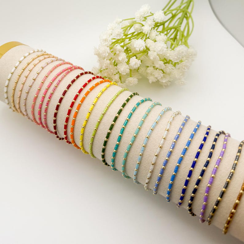 May include: A collection of colorful beaded bracelets displayed on a cylindrical surface. The bracelets feature various colors, including white, pink, red, orange, yellow, green, blue, and purple, with gold-colored beads. White flowers are in the background.