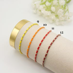 May include: Four beaded bracelets in yellow, orange, red, and burgundy. Each bracelet features small, colorful beads interspersed with gold-colored beads. The bracelets are displayed on a beige, cylindrical surface.