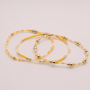 May include: Three circular bracelets made of small yellow and white seed beads interspersed with polished gold-tone metallic beads. The bracelets feature a repeating pattern of vibrant yellow, soft white, and reflective gold spherical accents.