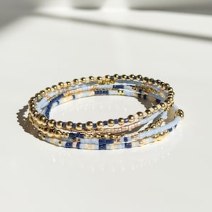 以下が含まれることがあります： 金色のビーズと、水色、ネイビー、クリーム色のシードビーズを使った、マルチストランドのラップブレスレット。白い表面に巻かれており、そのレイヤードデザインとカラーパレットが際立っています。ジュエリーアイテムです。