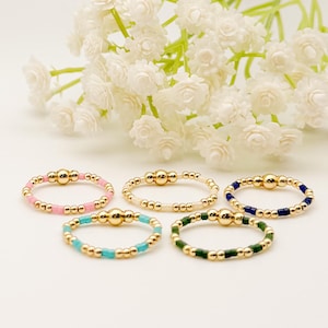 May include: Five beaded rings in various colors: pink, cream, turquoise, green, and navy blue. Each ring features a gold-colored central bead and smaller beads around the band. White flowers are in the background.