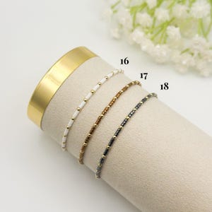 May include: Three delicate beaded bracelets in white, brown, and black, each with gold-colored accents. The bracelets are displayed on a beige surface, with the numbers 16, 17, and 18 visible. The background includes a gold-colored cylinder and white flowers.