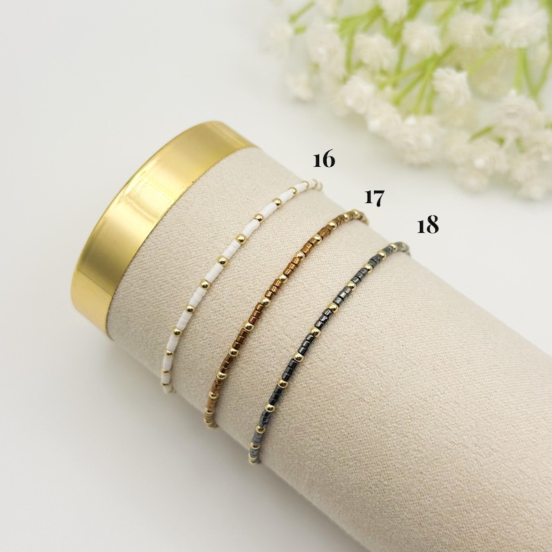 May include: Three delicate beaded bracelets in white, brown, and black, each with gold-colored accents. The bracelets are displayed on a beige surface, with the numbers 16, 17, and 18 visible. The background includes a gold-colored cylinder and white flowers.