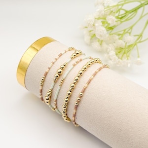 May include: A collection of gold beaded bracelets in various designs. Some bracelets feature small, colorful beads, while others have larger gold beads. The bracelets are displayed on a beige cylinder, with white flowers in the background.