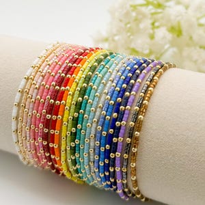 Könnte beinhalten: Eine Sammlung von Armbändern mit Perlen in Regenbogenfarben, jedes mit kleinen goldfarbenen Perlen. Die Armbänder werden auf einer weißen Oberfläche präsentiert und zeigen eine Vielzahl von Farbtönen von Weiß bis Braun.