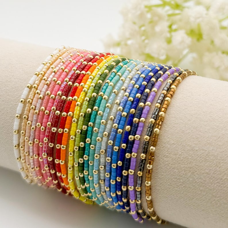 May include: A collection of beaded bracelets in a rainbow of colors, each with small gold-colored beads. The bracelets are displayed on a white surface, showcasing a variety of hues from white to brown.