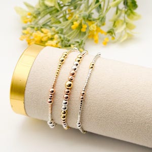 Puede incluir: Tres pulseras de cuentas en tonos dorados, plateados y oro rosa se exhiben sobre una superficie cilíndrica beige. Las pulseras presentan cuentas redondas de varios tamaños, creando un aspecto delicado y elegante. El fondo incluye flores amarillas.