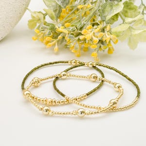 May include: Three gold beaded bracelets with varying bead sizes are displayed on a white surface. One bracelet has small olive green beads. Yellow flowers and green leaves are in the background, adding a touch of colour.