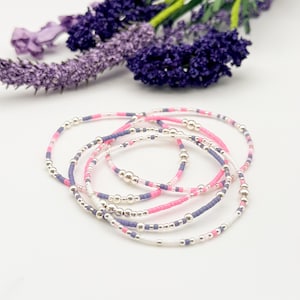 May include: Stack of beaded bracelets in pink, white, and purple with silver accents. The bracelets are arranged on a white surface, with a sprig of purple flowers in the background. The bracelets are delicate and feminine.
