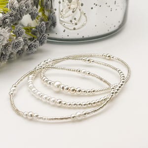 Puede incluir: Tres pulseras plateadas con detalles de perlas y cuentas. Las pulseras tienen un diseño delicado, combinando pequeñas cuentas con perlas más grandes. El estilo es sencillo y clásico, adecuado para el uso diario o ocasiones especiales.