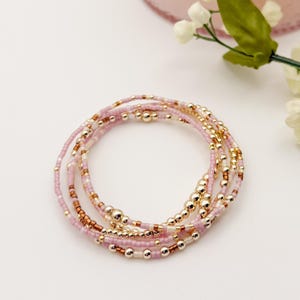 Puede incluir: Un conjunto de delicadas pulseras de cuentas en tonos rosa, dorado y transparente. Las pulseras están hechas con pequeñas cuentas redondas y cuentas doradas más grandes. Las pulseras están dispuestas en forma circular sobre una superficie blanca.