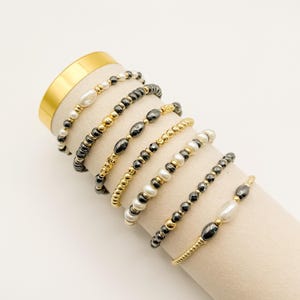 May include: A collection of beaded bracelets in various designs. The bracelets feature gold, black, and white beads, including pearls and hematite. Some bracelets have a mix of bead types, while others are solely gold or black. The bracelets are displayed on a beige surface.