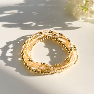 May include: A set of delicate gold and cream beaded bracelets stacked together. The bracelets are made of small, round beads and are perfect for layering.