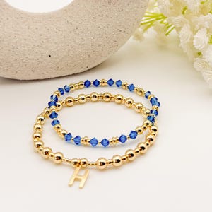 May include: Two gold beaded bracelets. One bracelet features blue crystal beads, and the other has a gold letter "H" charm. The bracelets are displayed on a white surface with a neutral background.