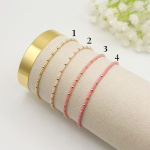 May include: Four delicate beaded bracelets in shades of beige, pink, and rose, accented with gold-colored beads. The bracelets are displayed on a neutral-colored cylindrical form, with a gold-colored band at the base. The bracelets are numbered 1 through 4.