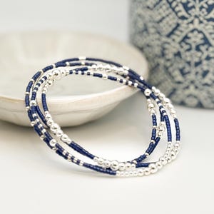 May include: Three beaded bracelets. The bracelets feature a combination of small, silver-colored beads and navy blue beads. The bracelets are coiled and rest on a white surface, with a white bowl and a blue and white patterned vase in the background.