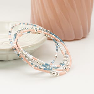 May include: A collection of beaded bracelets in shades of pink, blue, and silver. The bracelets are made with small, colorful beads and silver accents. The bracelets are displayed on a white surface with a pink vase in the background.