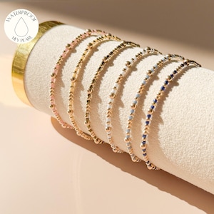 May include: A collection of delicate beaded bracelets in various colors, including pink, gold, white, light blue, and navy. The bracelets are displayed on a cream-colored cylindrical stand. The image includes a circular logo that says "WATERPROOF HEY PEARL."