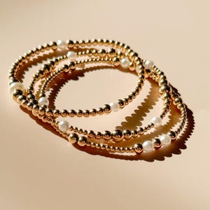 Puede incluir: Tres pulseras doradas con detalles de perlas blancas. Las pulseras están dispuestas en un patrón circular, con la luz reflejándose en las cuentas doradas. Las perlas están espaciadas uniformemente.