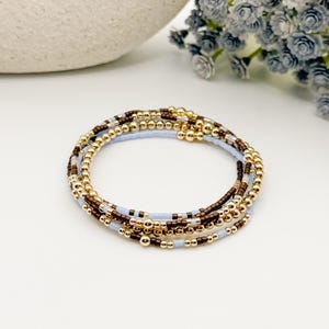 May include: A multi-strand beaded bracelet featuring a combination of metallic gold spheres, small dark brown cylindrical beads, and light blue cylindrical beads arranged in a repeating pattern to create a layered look on a circular elastic cord.