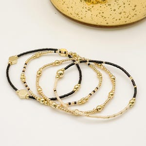 May include: Four beaded bracelets in gold, black, and cream colours. The bracelets feature small, round beads and larger, hexagonal and spherical gold-coloured accents. The bracelets are arranged on a white surface.