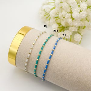 May include: Three beaded bracelets in white, teal, and blue with gold accents. The bracelets are displayed on a beige surface with a gold-colored cylinder in the background. The numbers 19, 20, and 21 are visible.