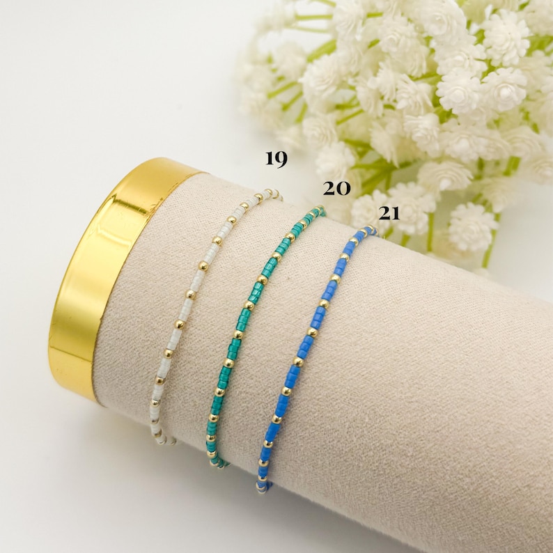 May include: Three beaded bracelets in white, teal, and blue with gold accents. The bracelets are displayed on a beige surface with a gold-colored cylinder in the background. The numbers 19, 20, and 21 are visible.