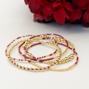 May include: A set of beaded bracelets in shades of gold, burgundy, and peach. The bracelets feature small, round beads and larger, gold-colored beads. The bracelets are arranged in a circular pattern, with a blurred red flower in the background.