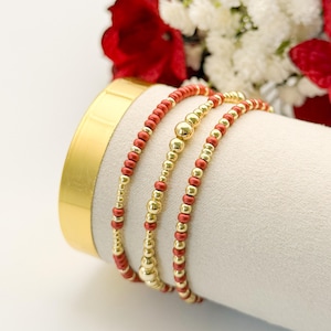 May include: Three gold and red beaded bracelets are displayed on a white surface. The bracelets have a mix of small red beads and larger gold beads. The background includes red and white flowers.