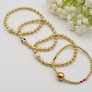 May include: Gold beaded bracelets with sports-themed charms. One bracelet features a volleyball charm, another a soccer ball, and a third a baseball. The baseball bracelet has red accents. White flowers are in the background.