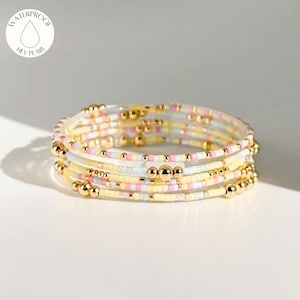 May include: A set of colourful beaded bracelets. The bracelets feature yellow, pink, white, and gold beads. The bracelets are wrapped around each other. The bracelets are waterproof.