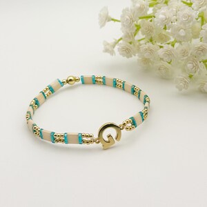 Puede incluir: Una pulsera de cuentas con cierre y detalles dorados. La pulsera presenta cuentas rectangulares color crema, cuentas cuadradas turquesas y pequeñas cuentas doradas. El cierre tiene forma de letra "G".