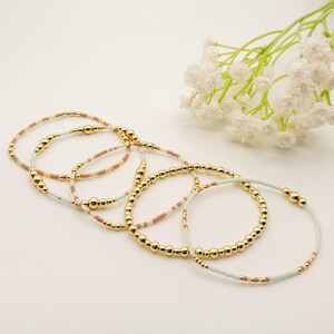 May include: Several gold beaded bracelets with light blue, tan, and gold bead accents. The bracelets are arranged overlapping, with a sprig of white flowers in the background.