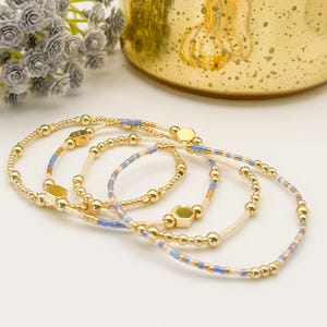 May include: Four gold beaded bracelets with blue and white accents. Each bracelet features small gold beads and hexagonal gold charms. The bracelets are arranged on a white surface, with a blurred gold background.