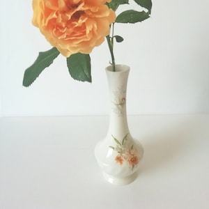 May include: A single peach-coloured rose with green leaves in a white ceramic vase with a floral pattern.