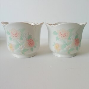 May include: Two white ceramic flower pots with a scalloped rim and floral design. The pots feature pink, yellow, and green flowers on a white background.